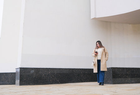 brunette woman wearing coat walking in the street along the large building, minimal urban background using smartphoneの写真素材