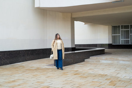 brunette woman wearing coat walking in the street along the large building, minimal urban backgroundの写真素材