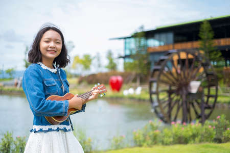 Portrait asian young girl wear jean jacket and white skirt playing ukulele at park.