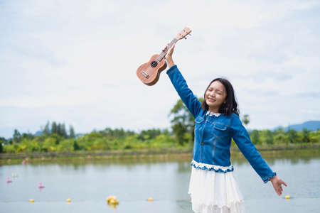 Portrait asian young girl wear jean jacket and white skirt playing ukulele at park.の写真素材