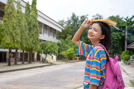 Asian young girl wear colorful shirt put book on her head standing at school.の写真素材