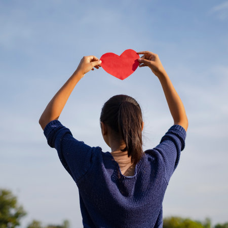 Girl hold red paper heart on head with blue sky background.