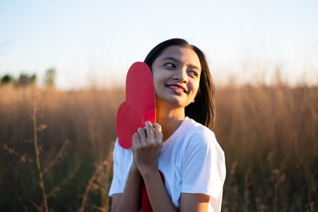 Happy Asian girl hold a red paper heart at brown grass sunset time.の写真素材