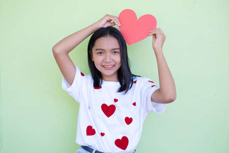 Happy Asian young girl holds red paper heart on green background.の写真素材