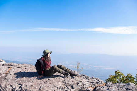 Tourist girl with backpack siting on the stone at the mountain, wear hat and looking panoramic view at Pha Mak Dook Phukradung National park at Thailand.の写真素材