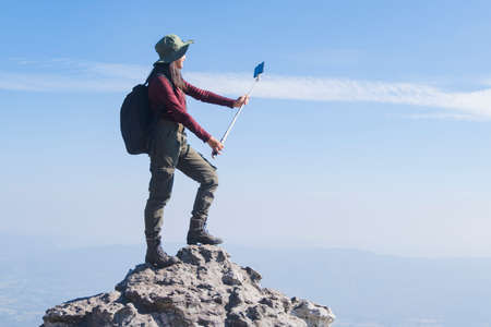 Tourist girl with backpack standing on the stone at the mountain use smart phone taking photo, wear hat and looking panoramic view at Pha Mak Dook Phukradung National park at Thailand.の写真素材