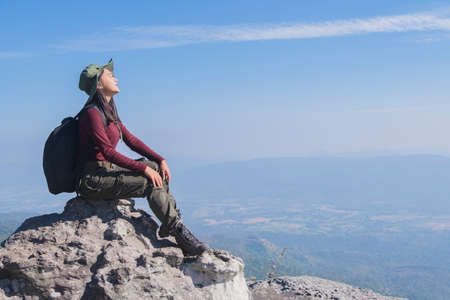 Tourist girl with backpack siting on the stone at the mountain, wear hat and looking panoramic view at Pha Mak Dook Phukradung National park at Thailand.の写真素材