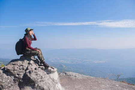 Tourist girl with backpack siting on the stone at the mountain, wear hat and looking panoramic view at Pha Mak Dook Phukradung National park at Thailand.の写真素材