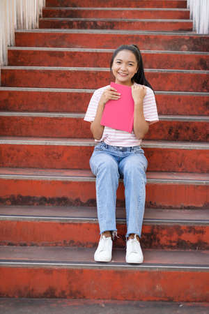 School girl  wear pink shirt and jean holding and holding pink book on stairs.の写真素材
