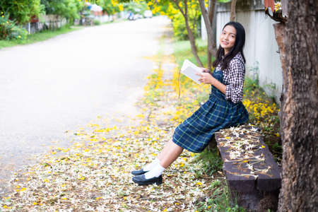 Young girl sitting on a bench reading a book under a beautiful tree.の写真素材