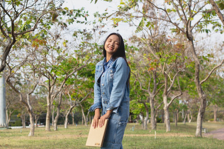 Cheerful young girl standing in the park, holding a book and smiling and wears jacket and jeans. lifestyle concept.の写真素材
