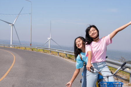 Two Young girl ride a bike on the road at the mountain with Wind Turbine, Khao Yai Thieng Electric Wind Turbine Thailand.の写真素材
