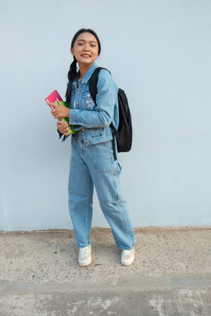 Student young girl standing at school wear jean hold book with backpack, blue background.の写真素材