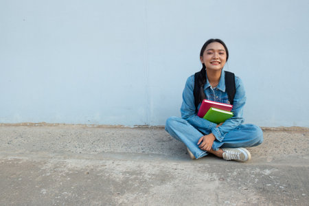 Student young girl sitting at school wear jean hold book with backpack, blue background.の写真素材