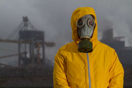 Man wearing gas mask standing infront of factory. Frontal shot. Background out of focus