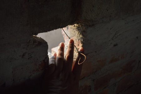 A hand reaches through a small hole in an old brick wall toward soft light, evoking curiosity, hope,...