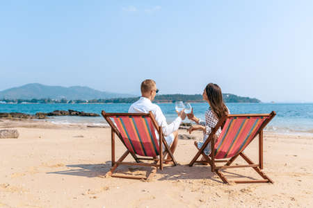 Smiling partners enjoying drinks champagne while resting on comfortable chairs by sea beach.の写真素材