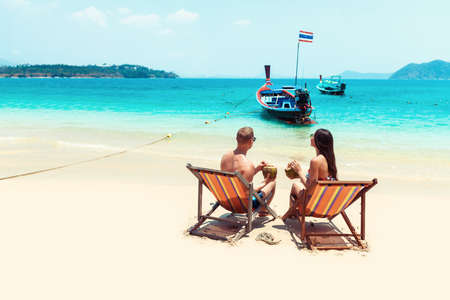 rear view of young couple with coconut cocktails relaxing in sun loungers on sandy beach.の写真素材