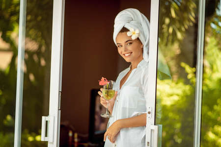 attractive woman in a white bathrobe and towel holding champagne glass and flower and smiling for the camera. Spaの写真素材