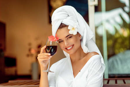 attractive woman in a white bathrobe and towel holding wine glass and flower and smiling for the camera. Spaの写真素材