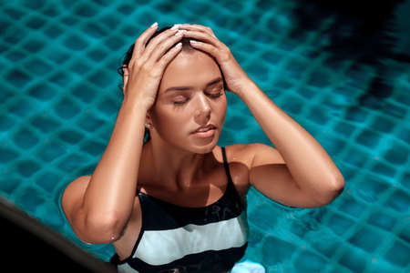 Tanned beauty girl in a fashion striped swimsuit lying in the pool water with closed eyes. Spa and relaxation concept. Summer vacationの写真素材