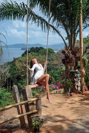 Charming girl in stylish white dress swinging on hanging swing in a tropical jungle. Concept of beauty, eleganceの写真素材