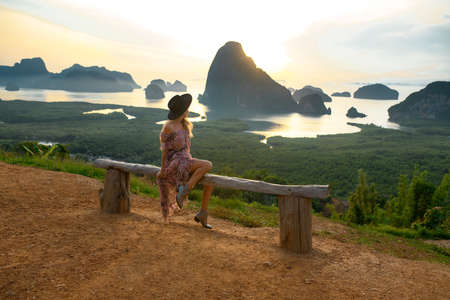 A pretty young girl in a hat and long dress is sitting on a wooden bench overlooking the lake and mountains. Boho style.の写真素材