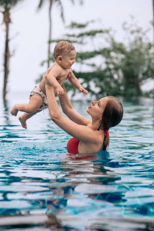 Mom is holding his baby son in his arms, while having fun in the luxurious pool. Family