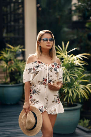 Stylish summer look. Attractive blonde with sunglasses in a short suit with floral prints and the hat in her hand posing for the camera.の写真素材