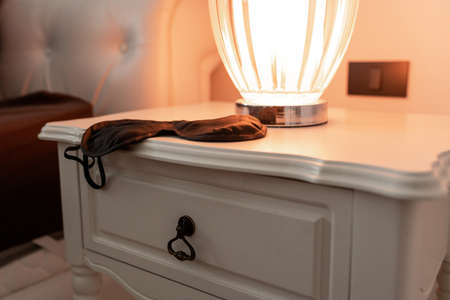 black sleeping mask lying on a white bedside table with a turned on table lampの写真素材