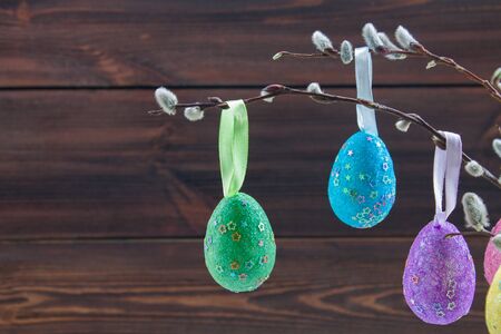 Multi-colored Easter eggs hanging from the branches of willow, wooden brown background
