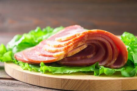 Sliced ham with fresh green lettuce leaves on a round cutting board. Meat products on a brown wooden table.の写真素材