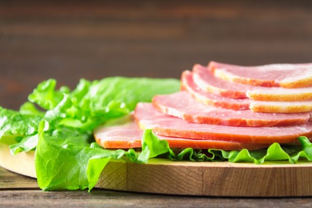 Sliced ham with fresh green lettuce leaves on a round cutting board. Meat products on a brown wooden table.の写真素材