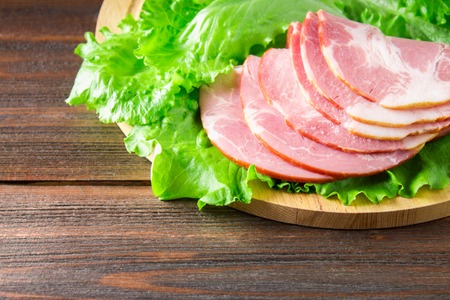 Sliced ham with fresh green lettuce leaves on a round cutting board. Meat products on a brown wooden table.の写真素材