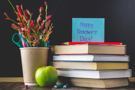 Concept of Teacher's Day. Objects on a chalkboard background. Books, green apple, plaque: Happy Teacher's Day, pencils and pens in a glass, sprig with autumn leaves.の写真素材