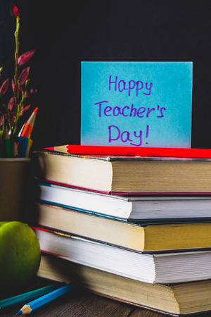 Concept of Teacher's Day. Objects on a chalkboard background. Books, green apple, plaque: Happy Teacher's Day, pencils and pens in a glass, sprig with autumn leaves.の写真素材