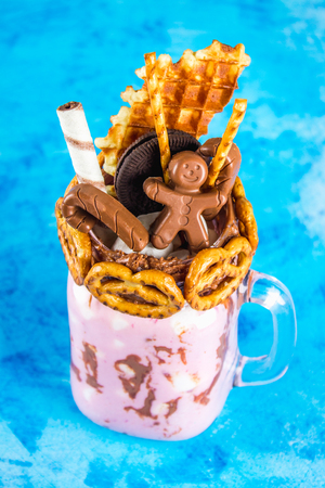 Freakshake from pink smoothie, cream. Monstershake with a chocolate man, cane, pretzels, biscuits, waffles and marshmelow. Extreme milkshake in a Mason jar. Blue concrete background.の写真素材