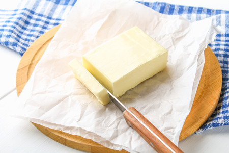 A bar of butter on a wooden board with a knife, on a white table. Ingredients for cookingの写真素材