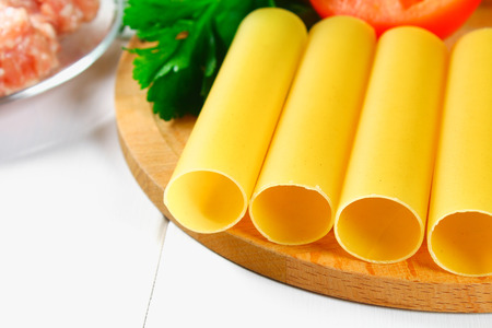 Italian pasta cannelloni. Raw tube for stuffing stuffing surrounded by ingredients for cooking, parmesan cheese, tomato, minced meat on a white wooden tableの写真素材