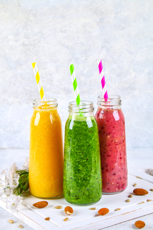 Green, yellow, purple smoothies in bottles of berries, greens, oranges on a white board on a gray tableの写真素材