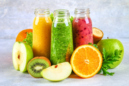 Green, yellow, purple smoothies in currant bottles, parsley, apple, kiwi, orange on a gray tableの写真素材