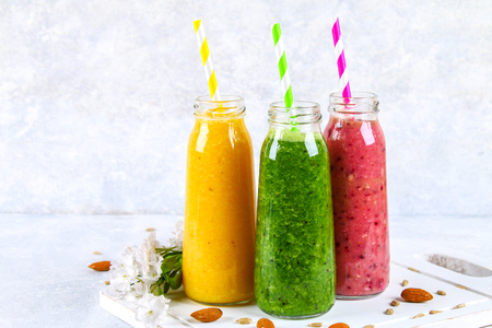 Green, yellow, purple smoothies in bottles of berries, greens, oranges on a white board on a gray tableの写真素材