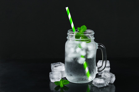 Ice water with ice and a mint leaf and a straw in a mason jar on a black background