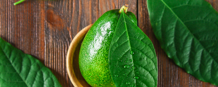A green raw ripe whole avocado fruit lies in a wooden bowl, surrounded by leaves on a brown tableの写真素材