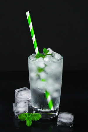 Ice water with ice and a leaf of mint and a straw in a glass on a black backgroundの写真素材
