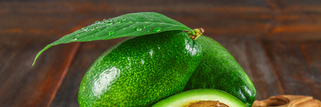 Three green raw ripe avocado fruits and a cut half with a bone with leaves on a wooden cutting board...