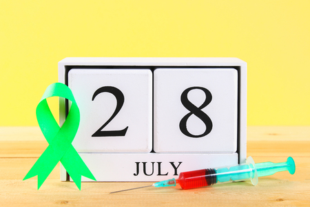 World Hepatitis Day. A wooden white calendar showing June 28 on a wooden table against a yellow wall background. Red jade and syringe with bloodの写真素材