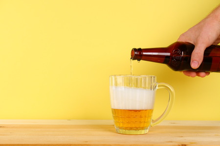 A man pours beer into a mug from a bottle on a yellow background on a wooden table. Copy spaceの写真素材