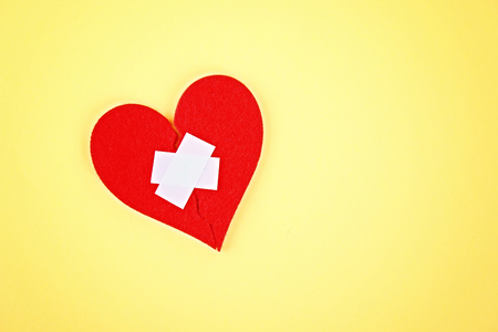 A red felt heart broken into two halves, glued together by a plaster on a yellow backgroundの写真素材