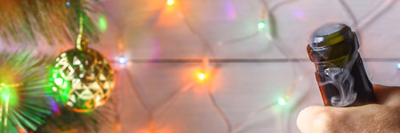 Men's hands open a bottle of champagne on the background of a New Year tree and garlands and glasses